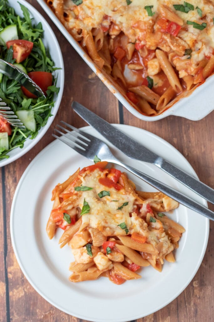 Overhead view of a plate of chicken and halloumi pasta bake with a knife and fork. Above rest of the pasta bake and a dish of green salad.