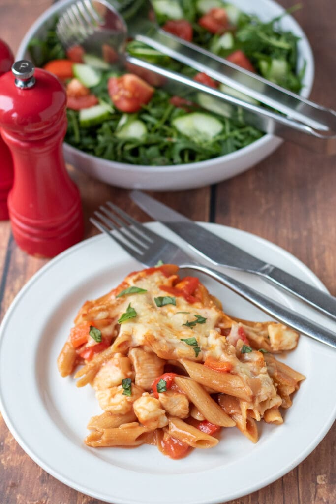 A portion of chicken halloumi pasta bake served on a plate with a knife and fork to the side. Salt and pepper cellars and a salad in a white serving bowl above.