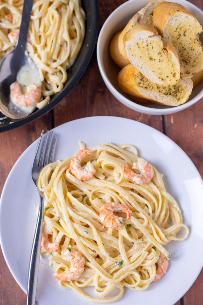 Overhead view of a plate of healthy creamy prawn lunguine with a fork to the right. Above rest of recipe in a pan and a bowl of garlic bread.
