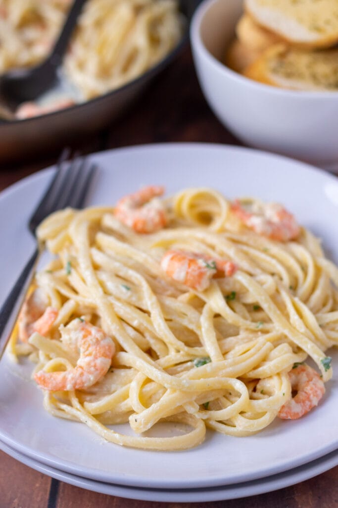 Creamy prawn pasta served on a plate garnished with chopped parsley. Fork to the side. Rest of dish in a pan and a bowl of sliced garlic bread at the top.