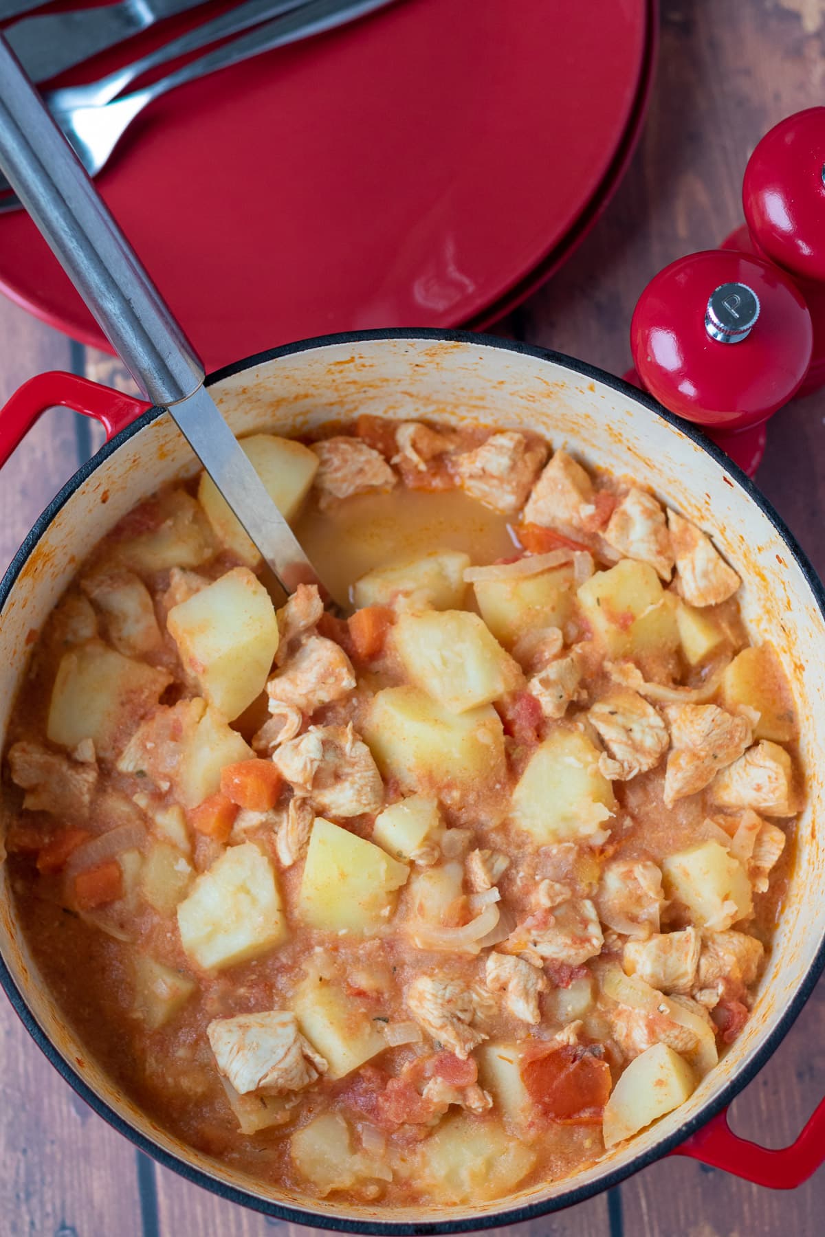 Overhead view of a casserole pot filled with chicken stew and tomatoes and potatoes. With a serving spoon in ready to serve.