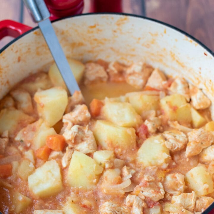 Chicken stew with tomatoes and potatoes cooked in a large casserole pot with a serving spoon in.