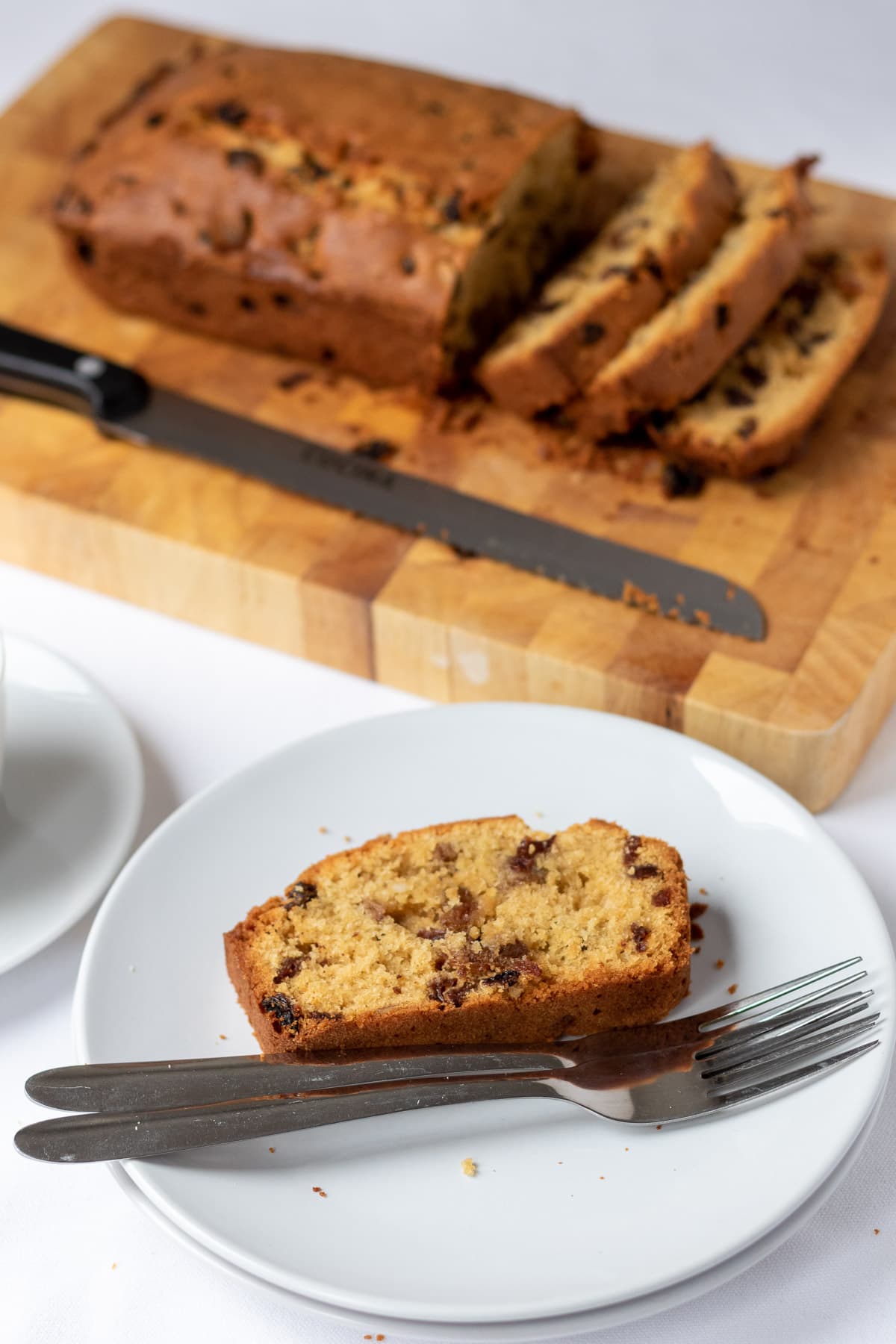 A slice of fruit loaf cake on a plate with two pastry forks alongside. Rest of loaf in the background on a chopping board.