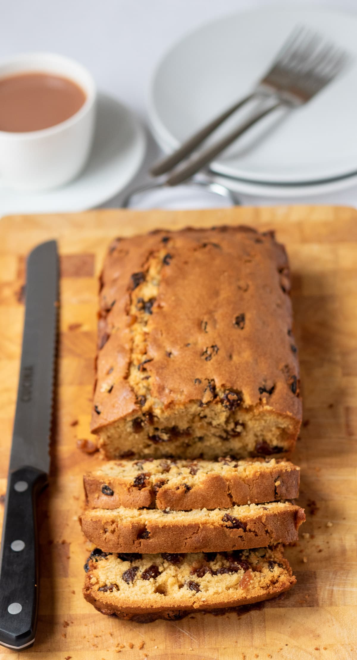 Fruit loaf cake on a chopping board. Three slices cut off the front. Cup of tea and serving plates behind.
