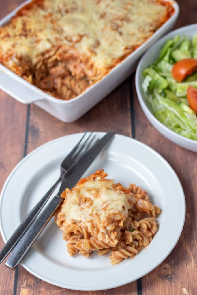 A portion of tuna tomato pasta bake served on a plate with a knife and fork to the side. Rest of the dish and a side salad above.