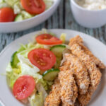 Healthy baked chicken strips served on a plate with a salad. Garlic mayonnaise in a ramekin and a bowl of salad in the background.