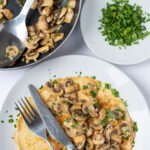 Overhead view of a plate with a savoury pancake with garlic mushrooms and a knife and fork to the side. Pan of garlic mushrooms above and dish of chopped parsley alongside.