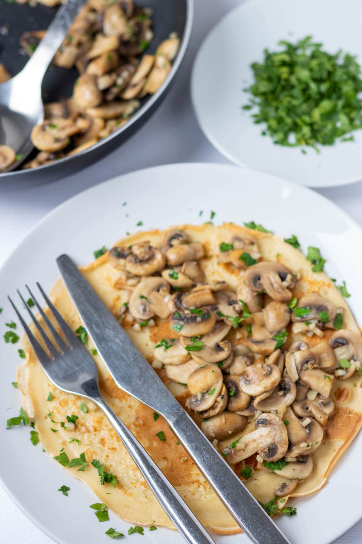 Savoury mushroom pancakes recipe served on a plate with a knife and fork. Dish of chopped parsley and frying pan of sauted mushrooms and garlic in the background.