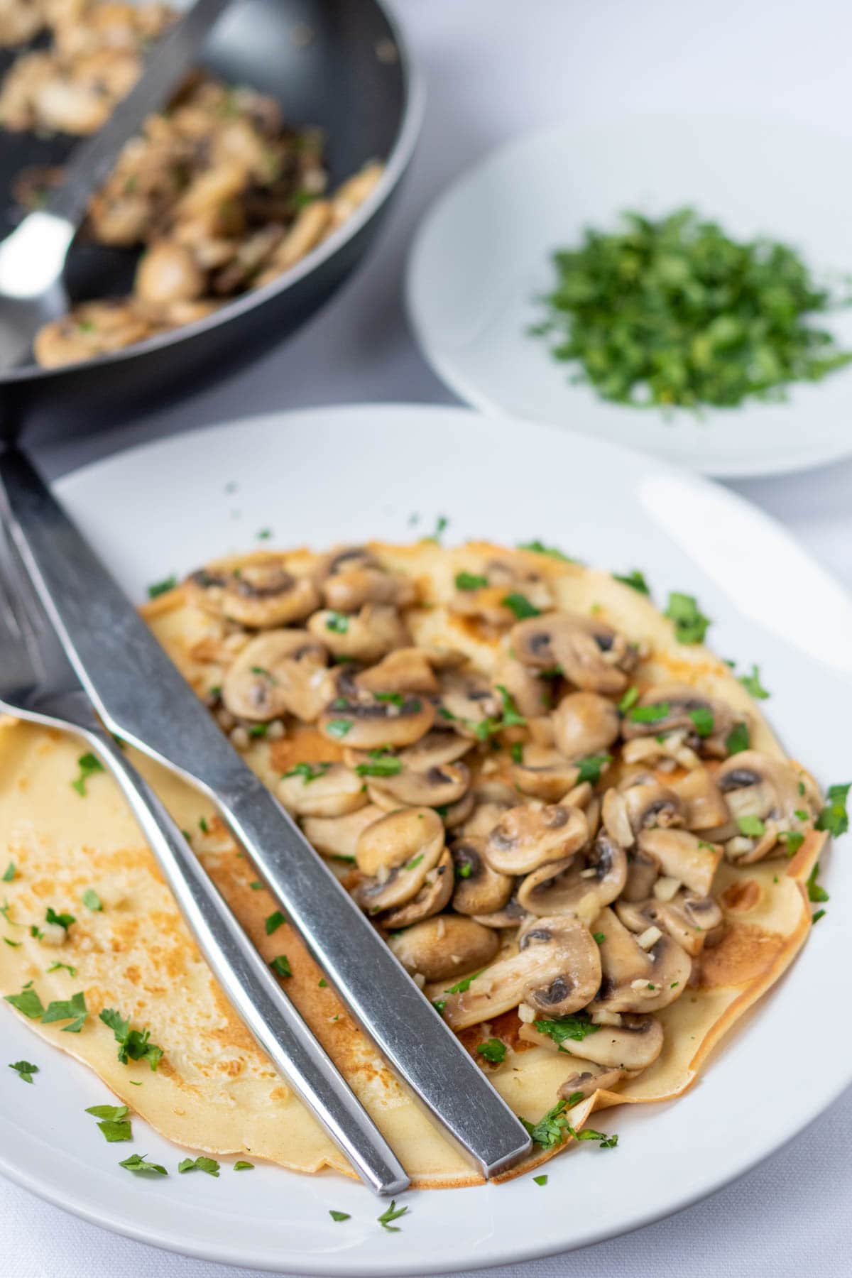 A savoury pancake with garlic mushrooms on a plate with a knife and fork to the side. Frying pan of garlic mushrooms and dish of chopped parsley in the background.