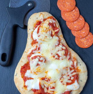 Easy pepperoni flatbread pizza cooked and on a black slate with a pizza cutter above and slices of pepperoni at the side.