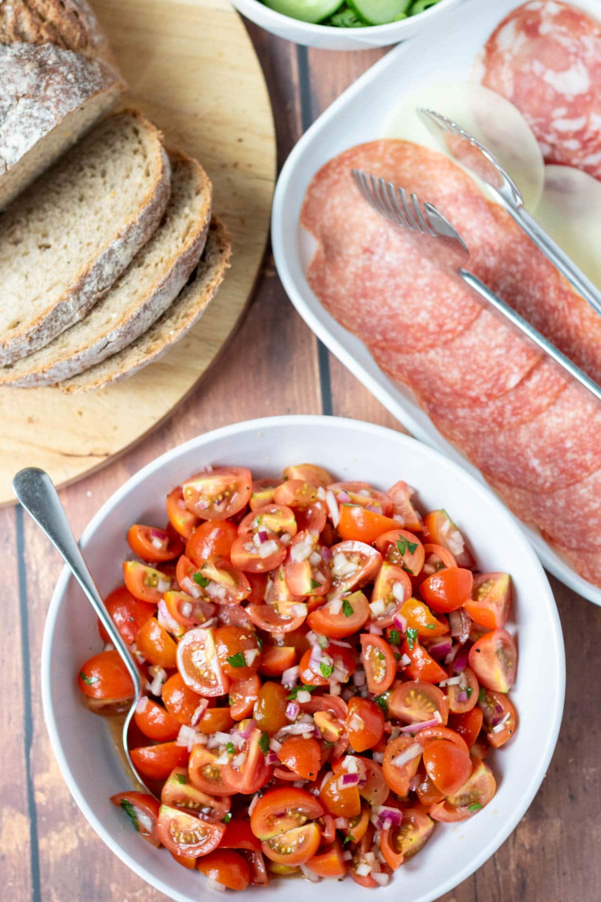 Overhead view of tomato onion salad served in a white bowl with a spoon in, alongside a platter of continental meats and a board of sliced crusty bread.