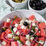 Watermelon and feta salad served in a large white serving dish with a serving spoon in. Two ramikins of green and black olives above.