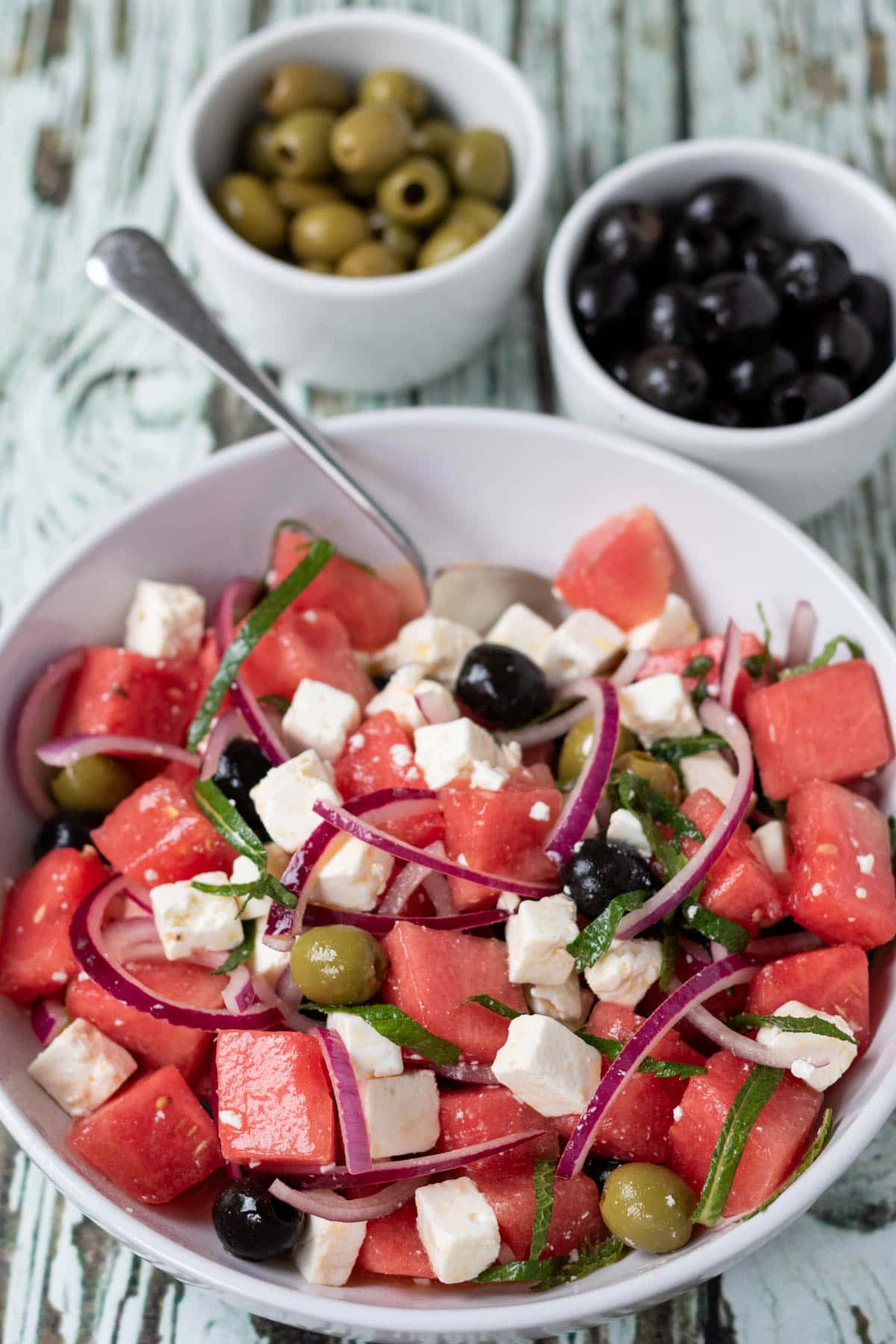Watermelon and feta salad served in a large white serving dish with a serving spoon in. Two ramikins of green and black olives above.