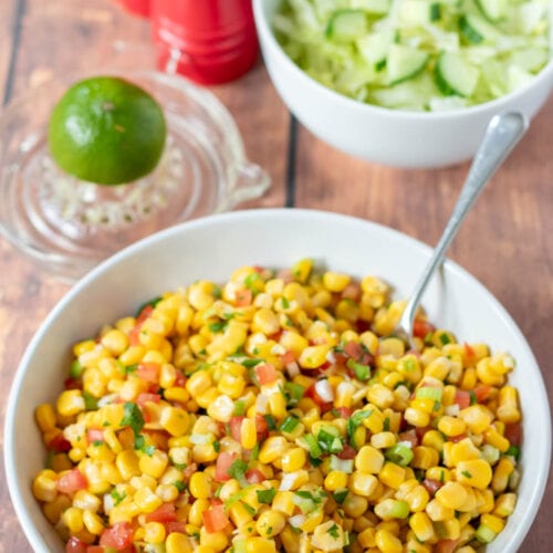 A bowl of quick corn salsa with a serving spoon in. Salad and a lemon juicer in the background.