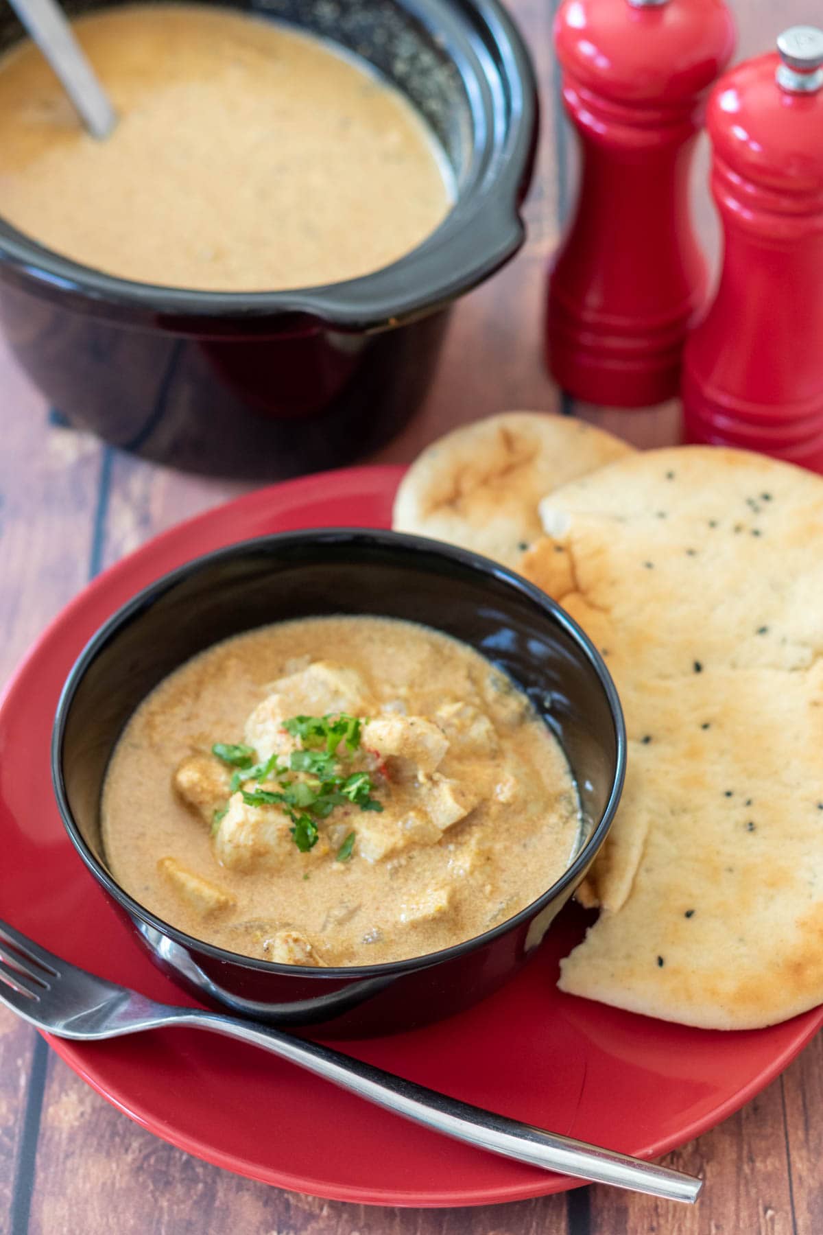 Easy slow cooker chicken curry served in a bowl garnished with coriander and a naan bread alongside. Slow cooker in the background.