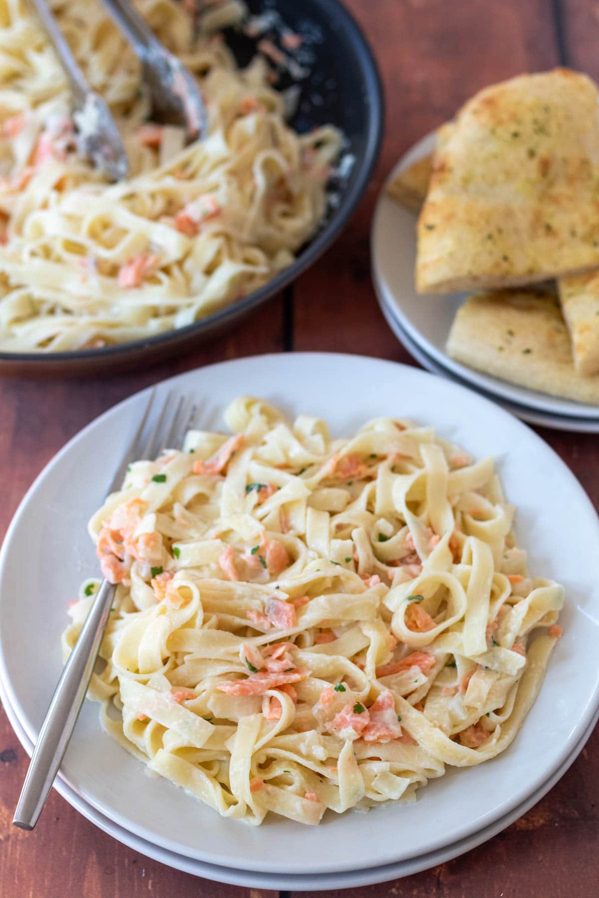 Healthy creamy salmon pasta served on a plate with a fork to the side. Rest of the dish in a pan behind alongside a plate of garlic bread.