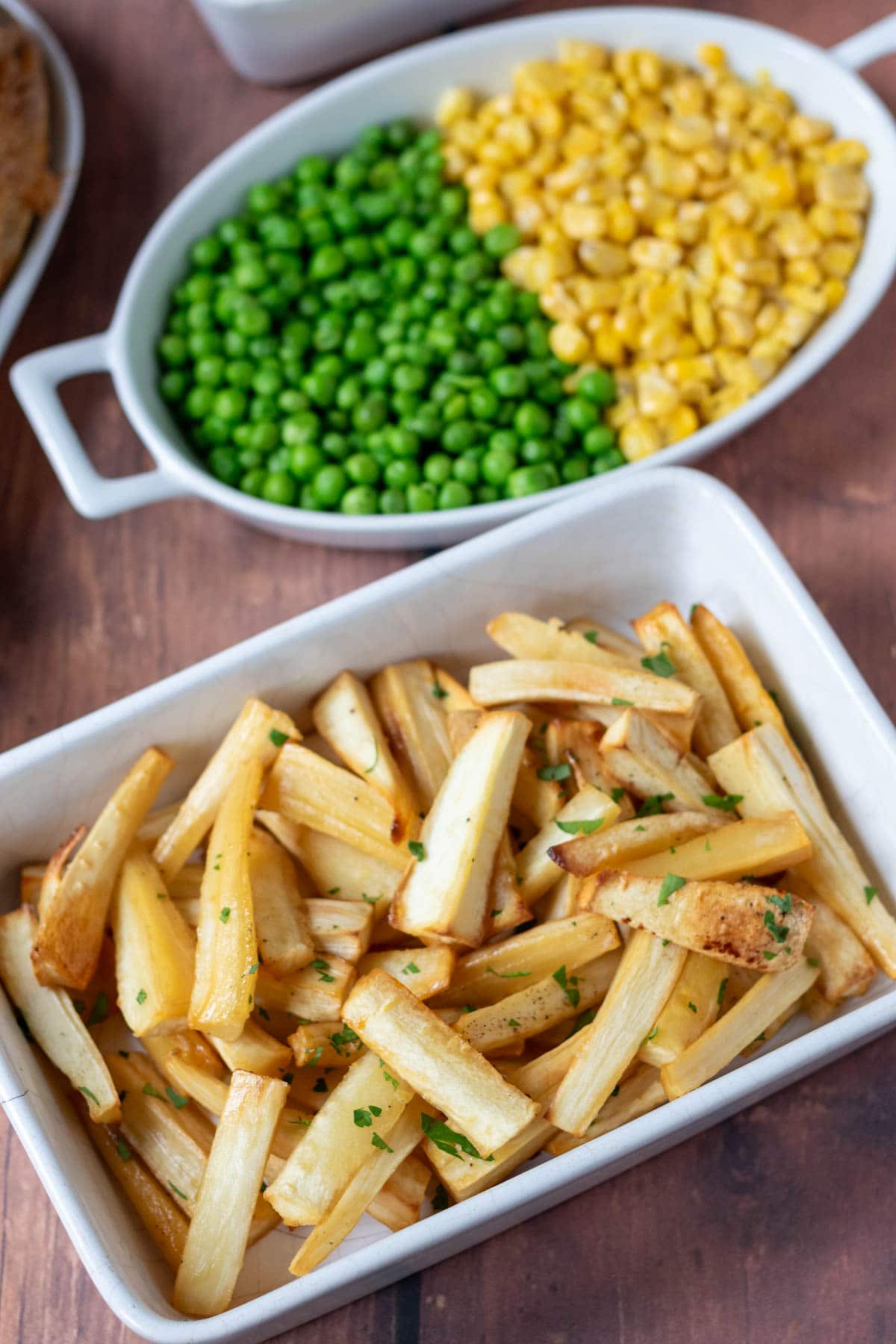 Honey glazed parsnips in a serving dish with a side dish of peas and sweetcorn above.