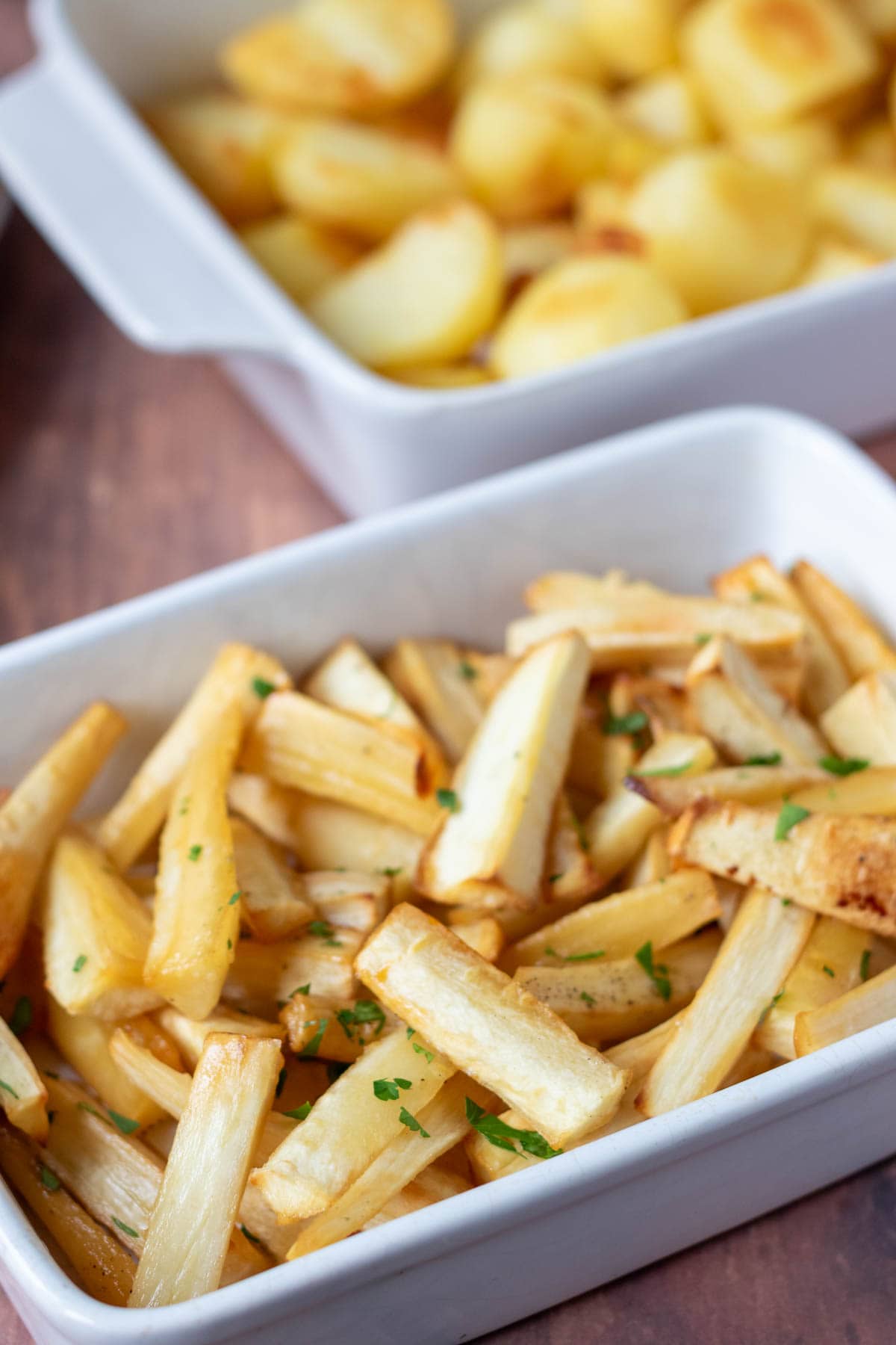 Roasted honey glazed parsnips in a serving dish with a dish of roast potatoes behind.