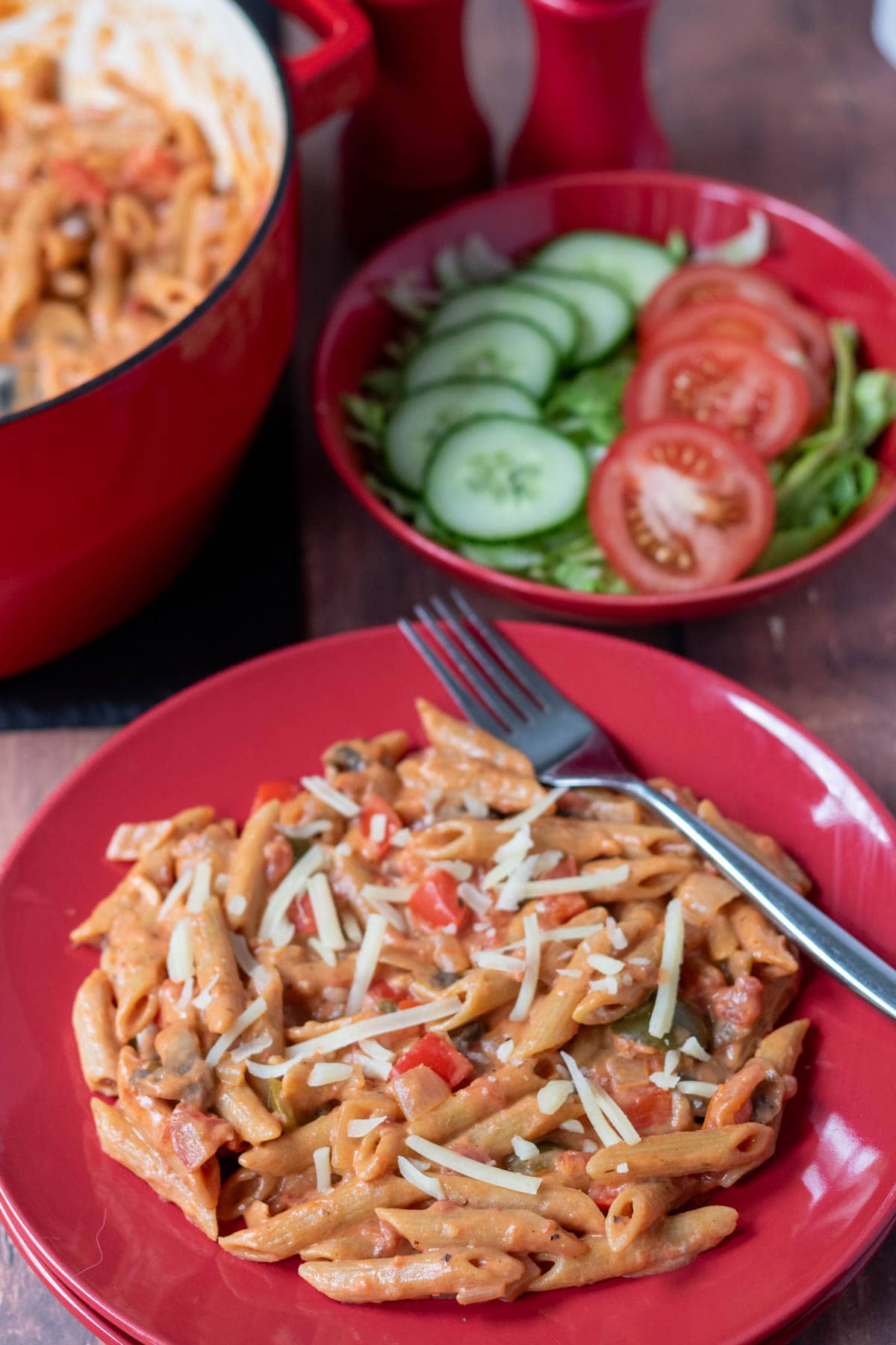 One pot vegetable pasta plated with a fork to the side. Bowl of lettuce, sliced tomato and cucumber in the background alongside a large casserole pot.