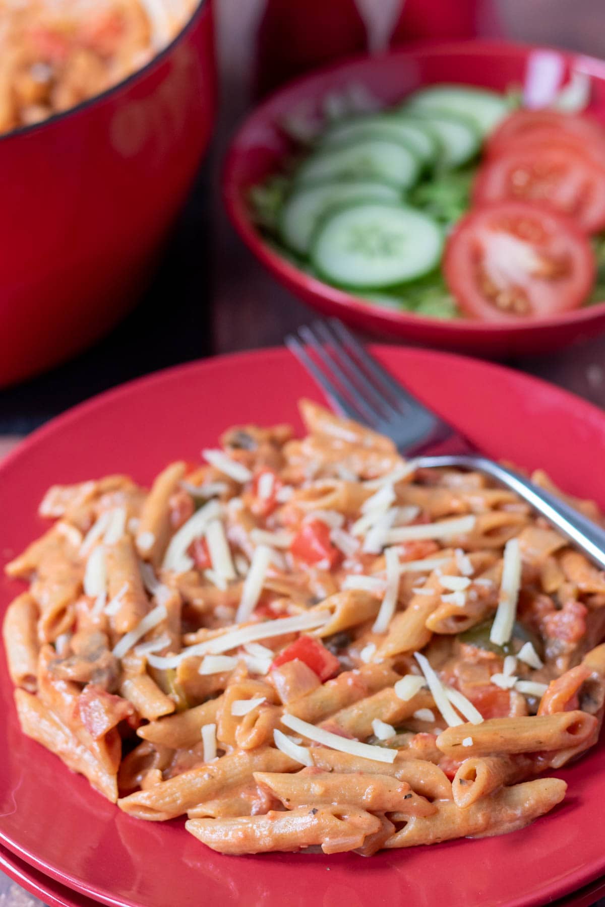 Close up of a plate of easy veggie pasta with a fork on the side. In the background a side salad and the cooking casserole pot.