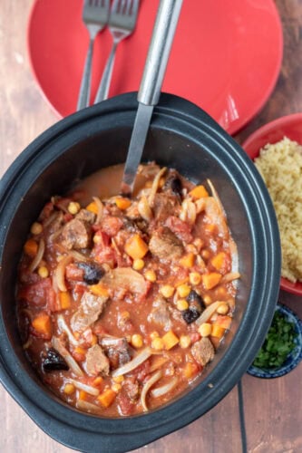 Easy slow cooker Moroccan lamb overhead view in slow cooker with serving spoon. Serving plates and forks at top and bowl of couscous to the right side.