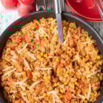 Overhead view of one pan chicken and quinoa cooked and in a large deep pan, ready to serve with a serving spoon in. Salt and pepper cellars and serving bowls with forks in above.