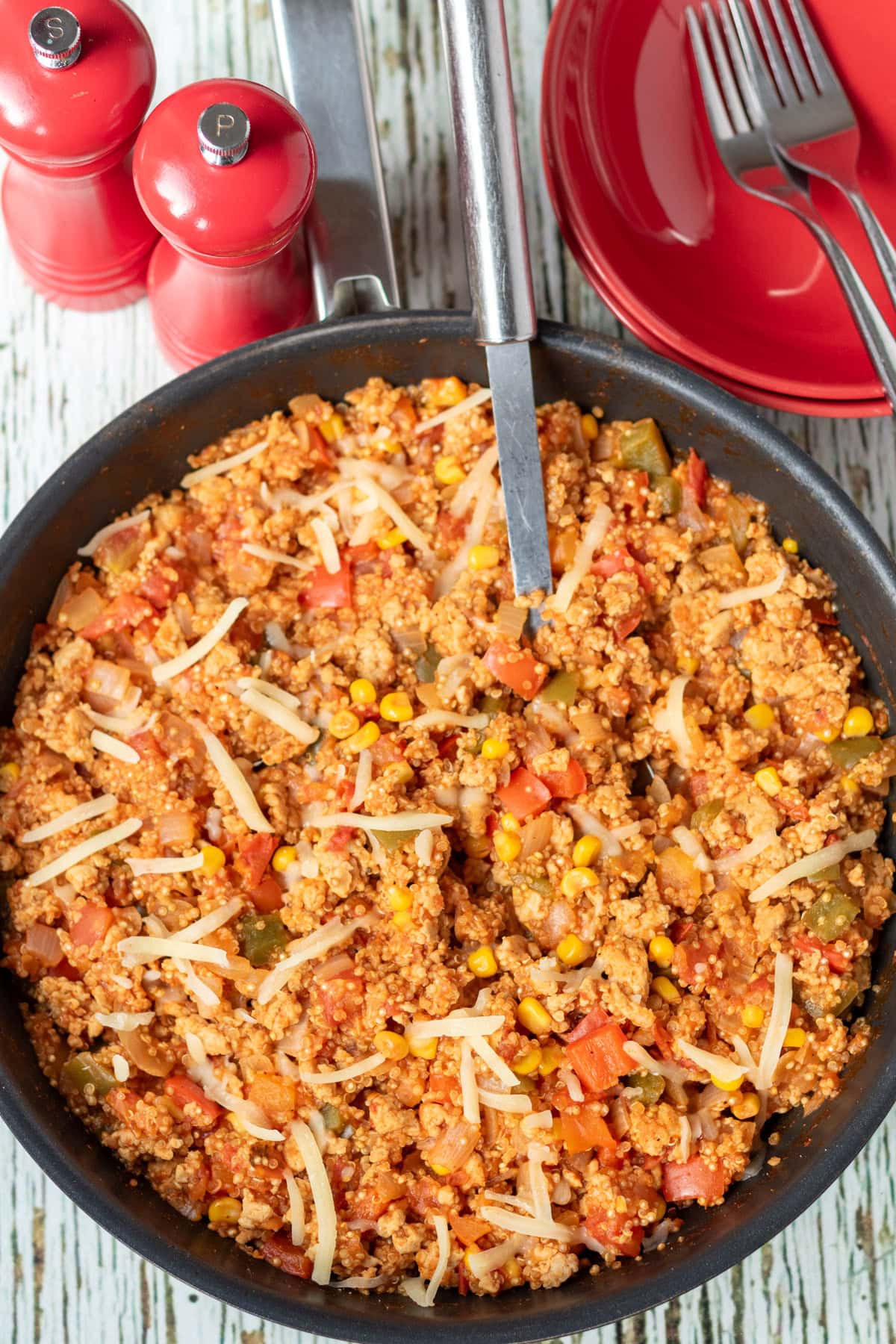 Overhead view of one pan chicken and quinoa cooked and in a large deep pan, ready to serve with a serving spoon in. Salt and pepper cellars and serving bowls with forks in above.
