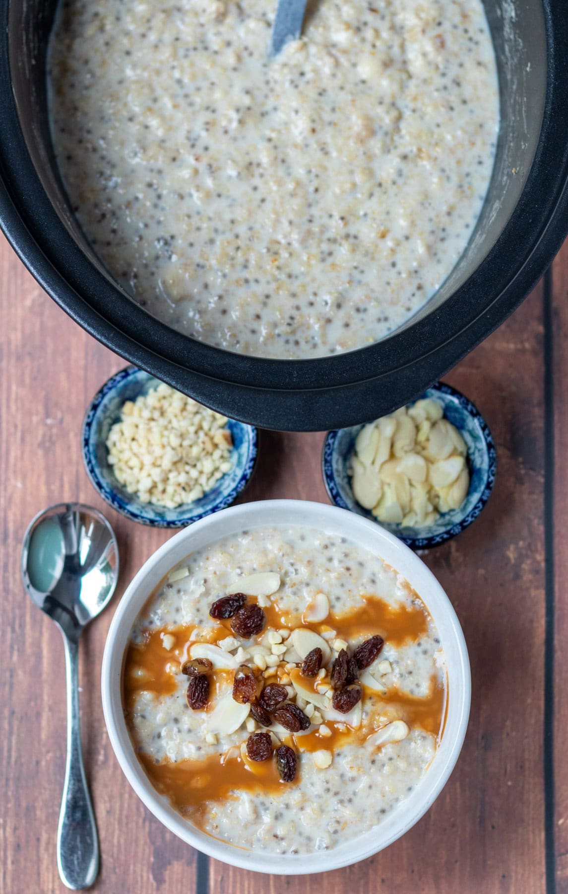 Overhead view of slow cooker with banana porridge in it at the top, at the bottom a bowl with a serving of slow cooker banana porridge in topped with sultana's, nuts and caramel topping. Breakfast spoon to the left and dishes of chopped nuts in between.