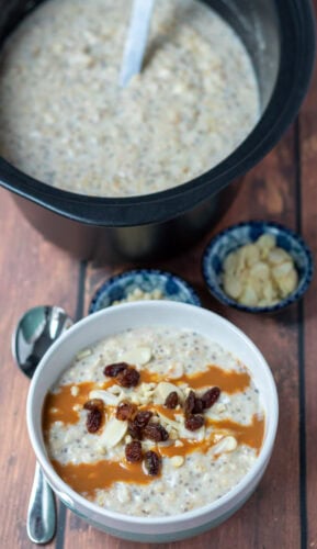 A bowl of slow cooker banana porridge topped with sultanas, nuts and caramel topping at the bottom. A spoon to the left. Rest of porridge at top in slow cooker. Dishes of chopped nuts in between.