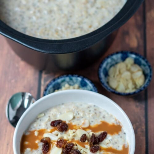 A bowl of slow cooker banana porridge topped with sultanas, nuts and caramel topping at the bottom. A spoon to the left. Rest of porridge at top in slow cooker. Dishes of chopped nuts in between.