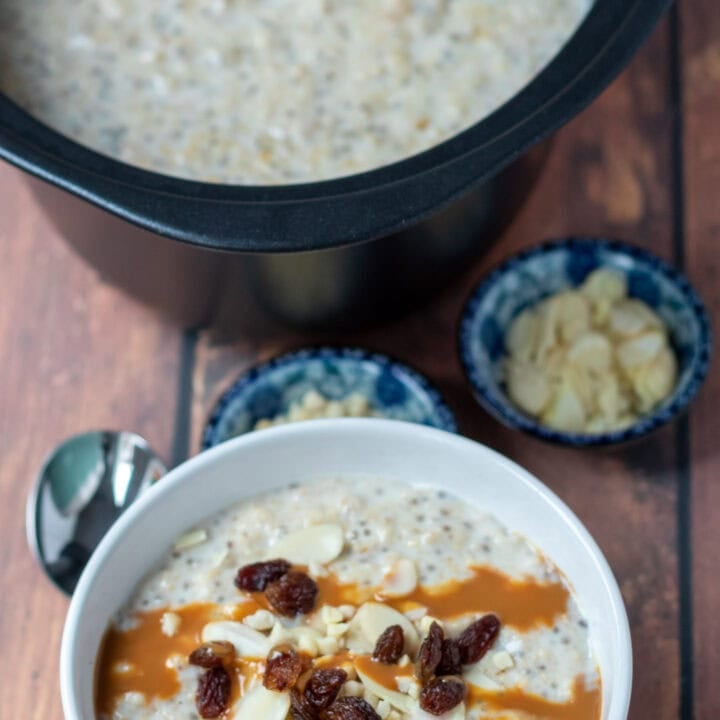 A bowl of slow cooker banana porridge topped with sultanas, nuts and caramel topping at the bottom. A spoon to the left. Rest of porridge at top in slow cooker. Dishes of chopped nuts in between.