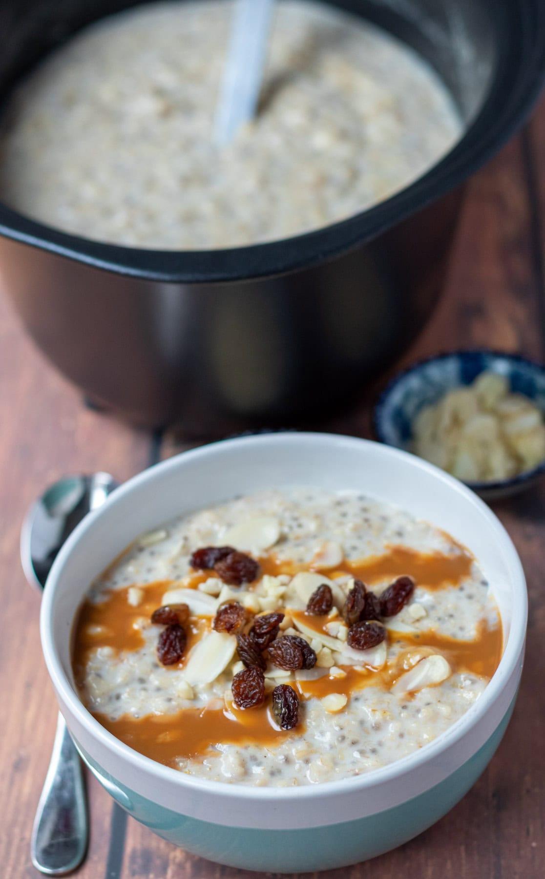 Slow cooker banana porridge in slow cooker container with a serving spoon in at top. Below an individual serving topped with sultanas, chopped nuts and caramel topping with a spoon to the left of the bowl.