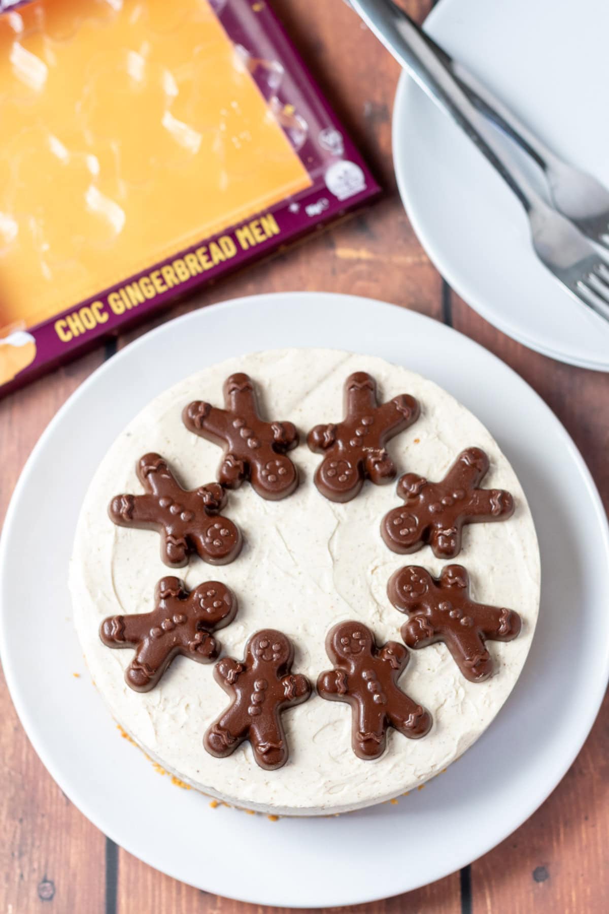 Overhead view of full no-bake gingerbread cheesecake on a plate. Above it to the left the empty packaging of the chocolate gingerbread biscuits and to the top right serving plates with dessert forks on.