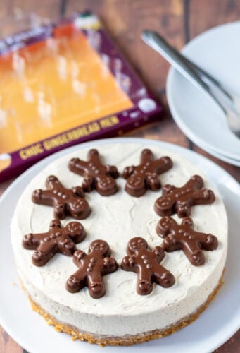 Easy no-bake gingerbread cheesecake on a plate decorated with chocolate gingerbread biscuits. In the background to the left the empty package that contained the chocolate gingerbread biscuits and to the right two plates with dessert forks on.