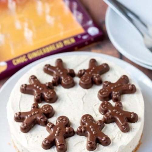 Easy no-bake gingerbread cheesecake on a plate decorated with chocolate gingerbread biscuits. In the background to the left the empty package that contained the chocolate gingerbread biscuits and to the right two plates with dessert forks on.