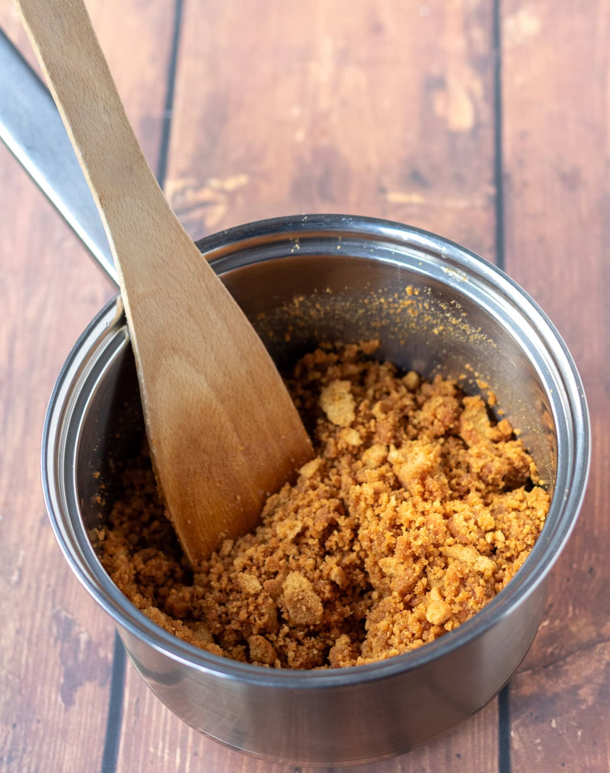 Crushed gingernut biscuits being stirred with a wooden spatula into melted butter in a saucepan.