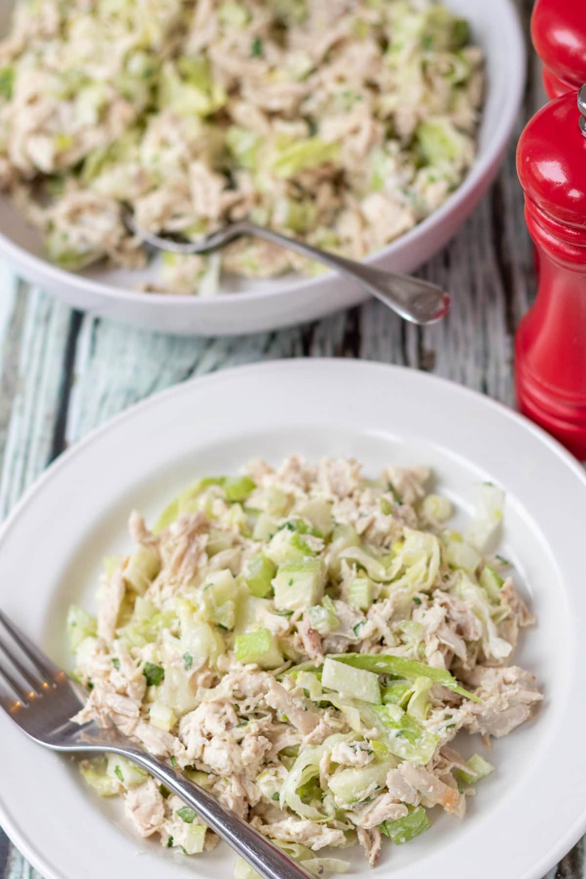 Overhead view of a serving dish of turkey salad at the top with a spoon in. Below it a serving of the salad on a plate with a fork to the left side. Salt and pepper cellars to the right in between the plate and serving bowl.