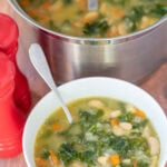 A bowl of white bean and kale soup with a soup spoon in. Behind is the rest of the soup in a large saucepan with a ladle in. To the left are salt and pepper cellars.