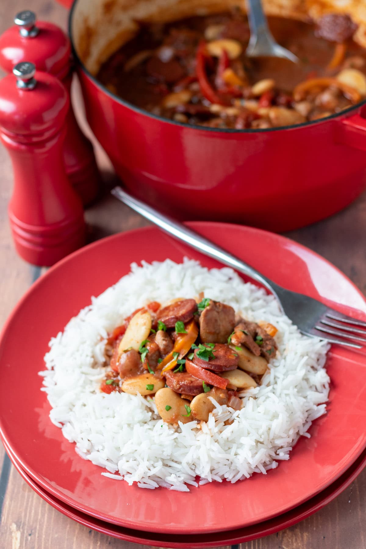 One pot spanish chicken and chorizo stew served on a plate on a bed of rice with a fork to the side. Above is rest of recipe in a large casserole pot to the left hand side are salt and pepper cellars.