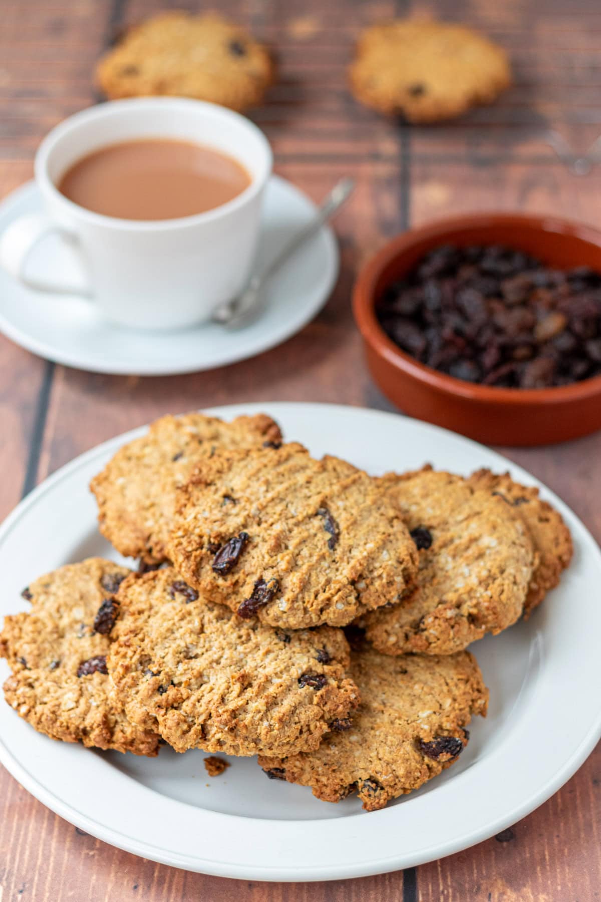 A batch of healthy raisin oat cookies on a plate, behind a cup of tea in a tea cup and a dish of raisins. Behind that a wire baking rack with other cookies cooling on it.