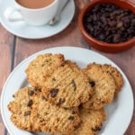 A plate stacked with raisin oat cookies at the font. Behind a cup of tea and a dish of raisins.