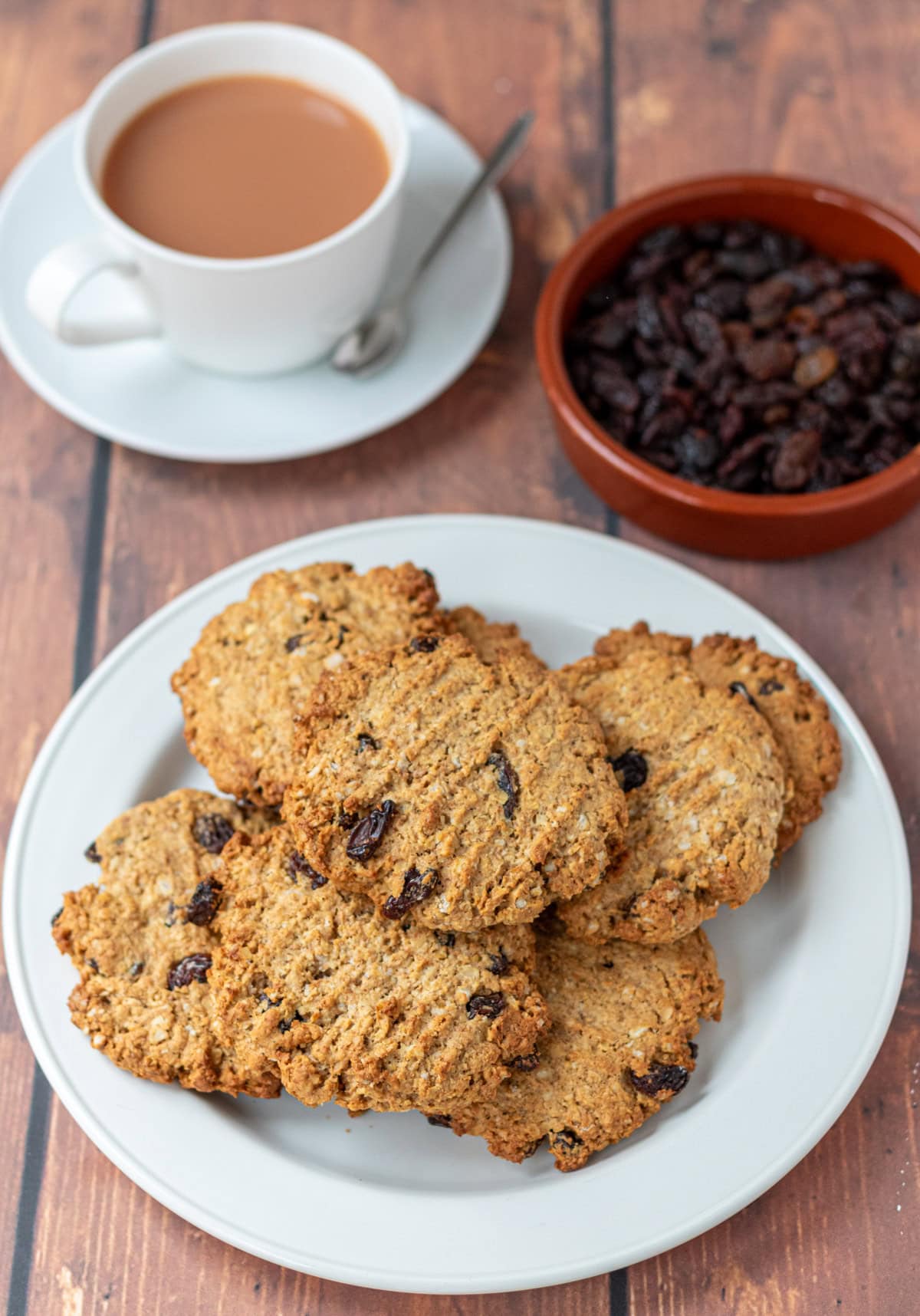 A plate stacked with raisin oat cookies at the font. Behind a cup of tea and a dish of raisins.