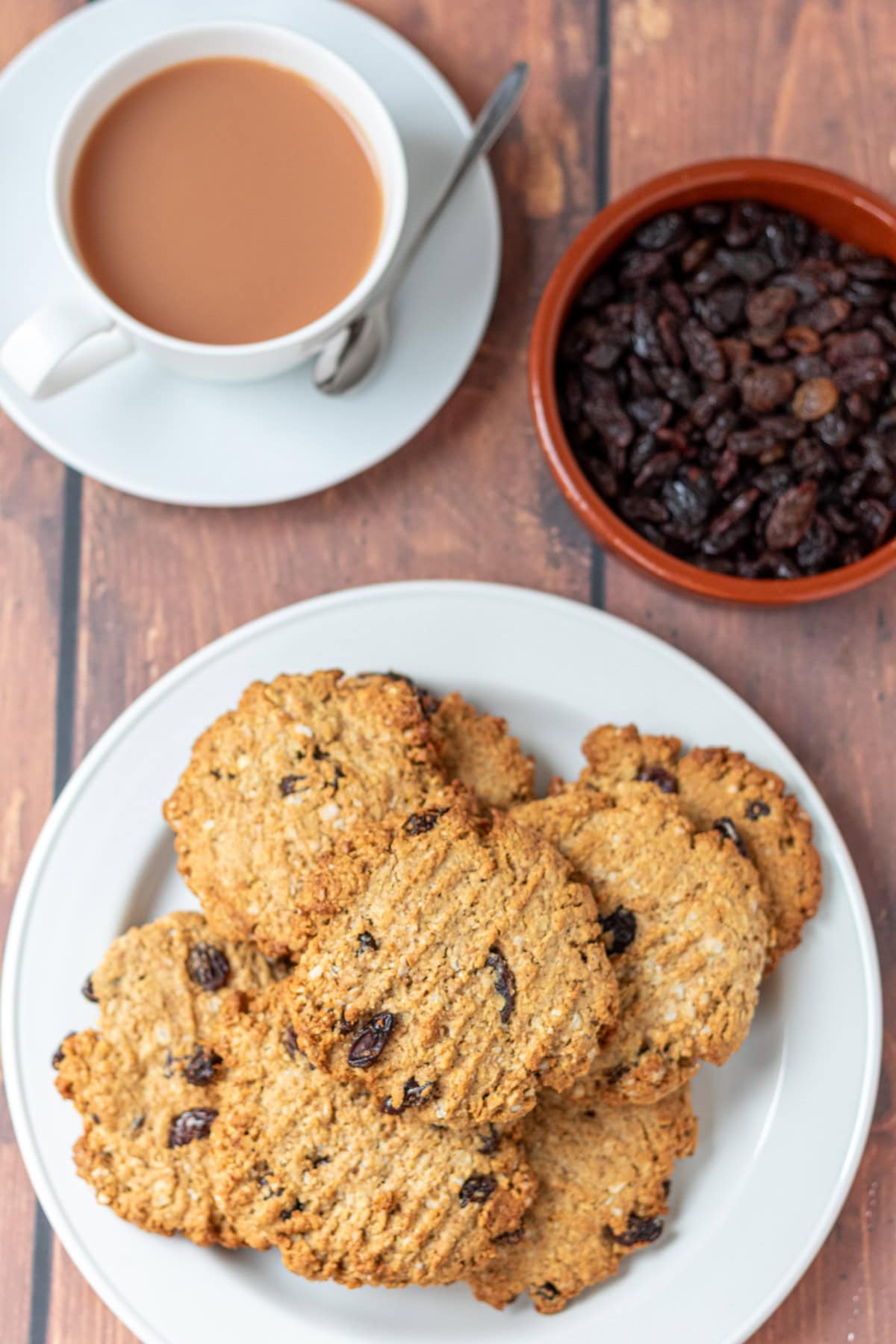 Overhead view of a plate of raisin oat cookies, stacked. Along side a cup of tea and a dish of raisins.
