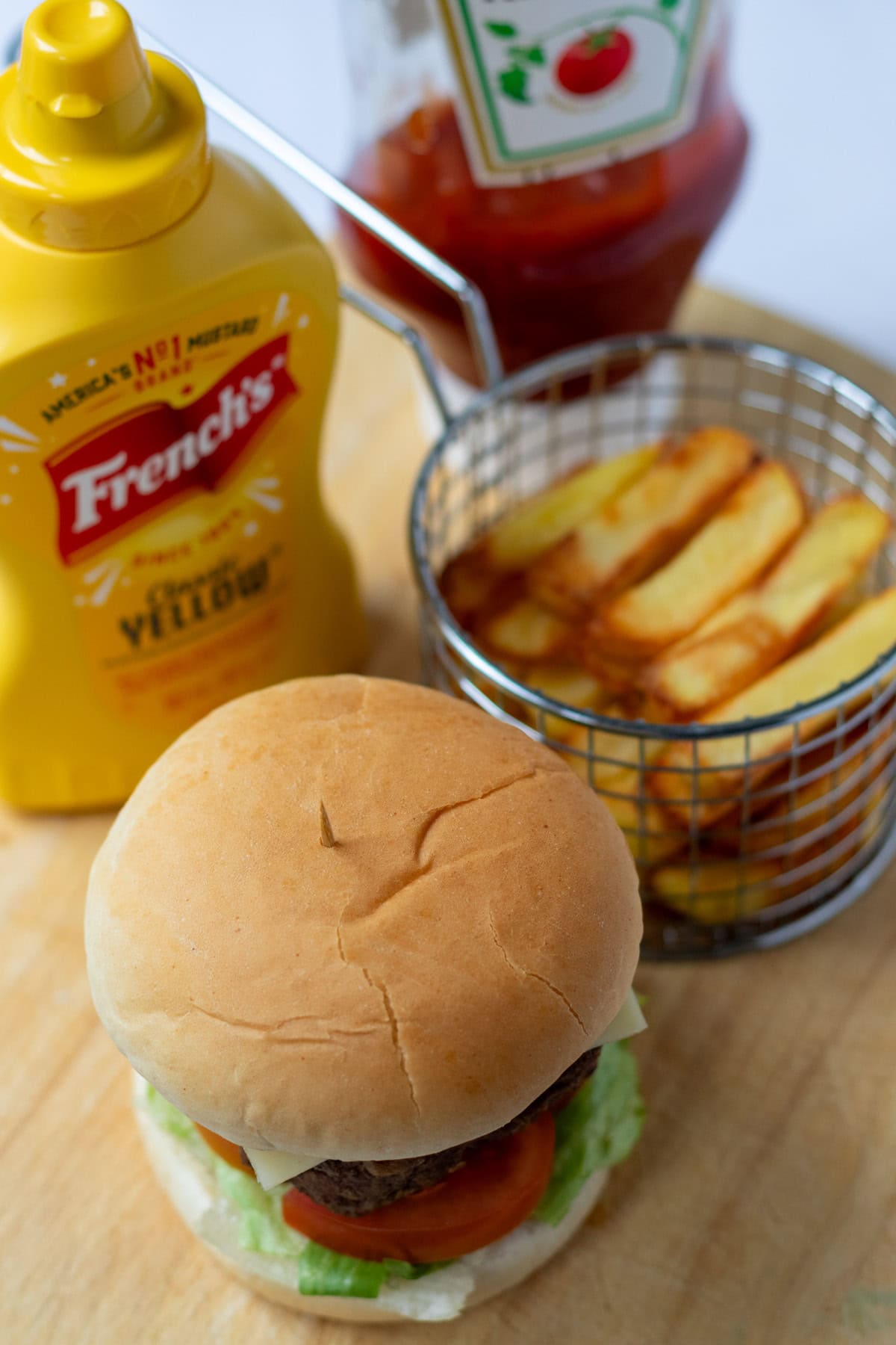 Overhead view of a lamb burger in a bun, french fries in a mini fryer basket above and mustard and tomato sauce condiments to the side.