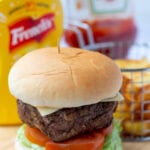 Easy simple lamb burger in a toasted burger bun on top of a bed of lettuce and sliced tomatoes. In the background mustard and ketchup plastic bottles and a basket of french fries.