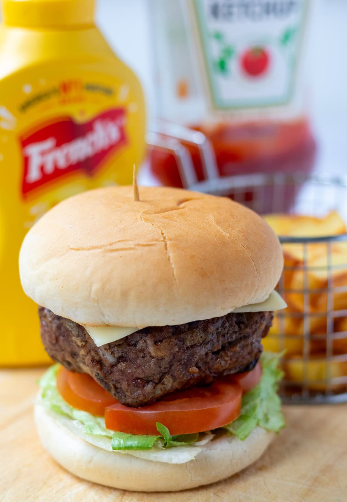Easy simple lamb burger in a toasted burger bun on top of a bed of lettuce and sliced tomatoes. In the background mustard and ketchup plastic bottles and a basket of french fries.