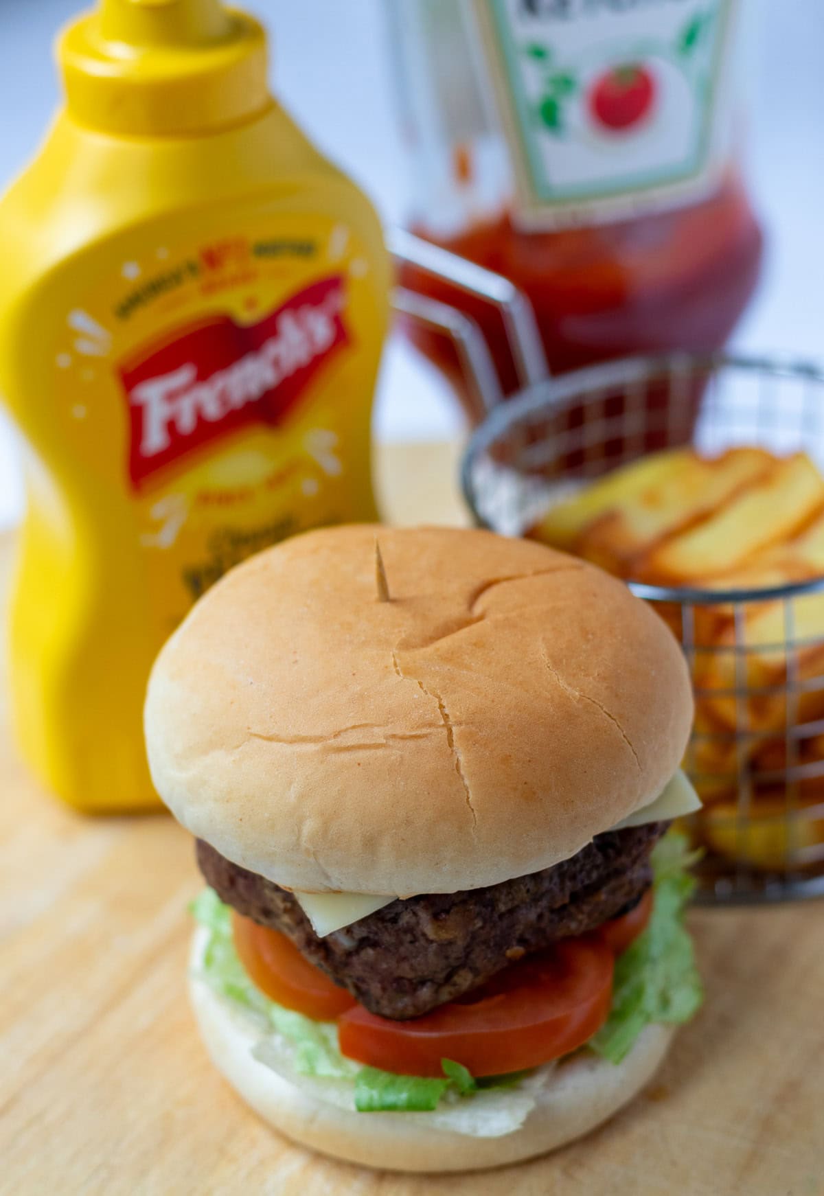 Lamb burger in a burger bun on a bed of lettace and sliced tomatoes. Alongside a basket of french fries. In the background a mustard bottle and a ketchup bottle.