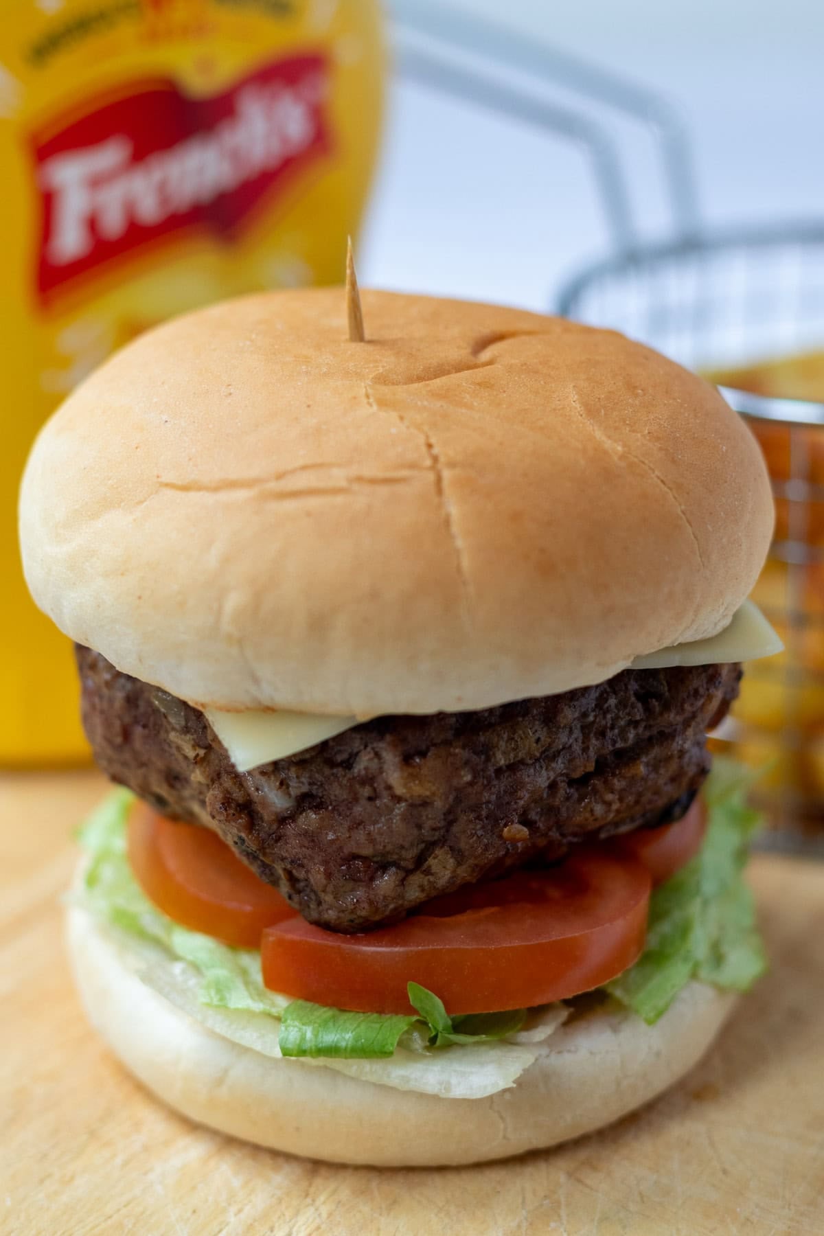 Homemade lamb burger in a burger bun with lettuce and sliced tomatoes. In the background a mustard bottle and a basket of french fries.