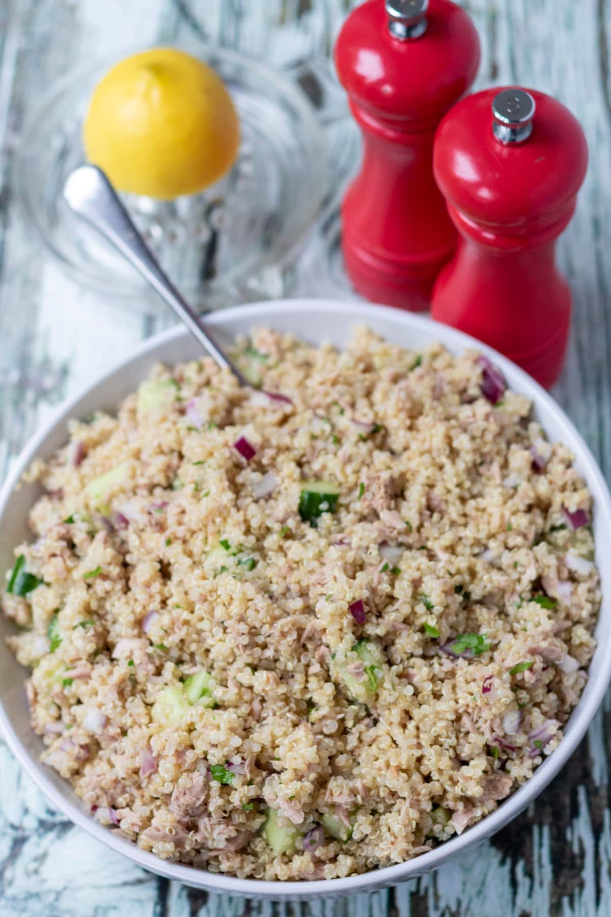 Bowl of quinoa tuna salad served with a serving spoon in. Lemon juicer and salt and pepper cellars behind.