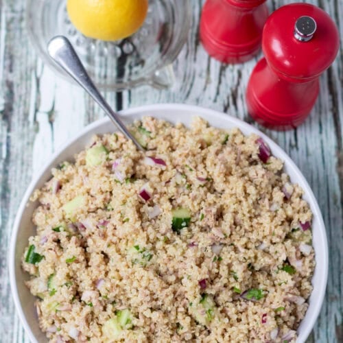 Overhead view of a serving dish of quick tuna quinoa salad with a serving spoon in. Above half a lemon in a lemon juicer and salt and pepper cellars.
